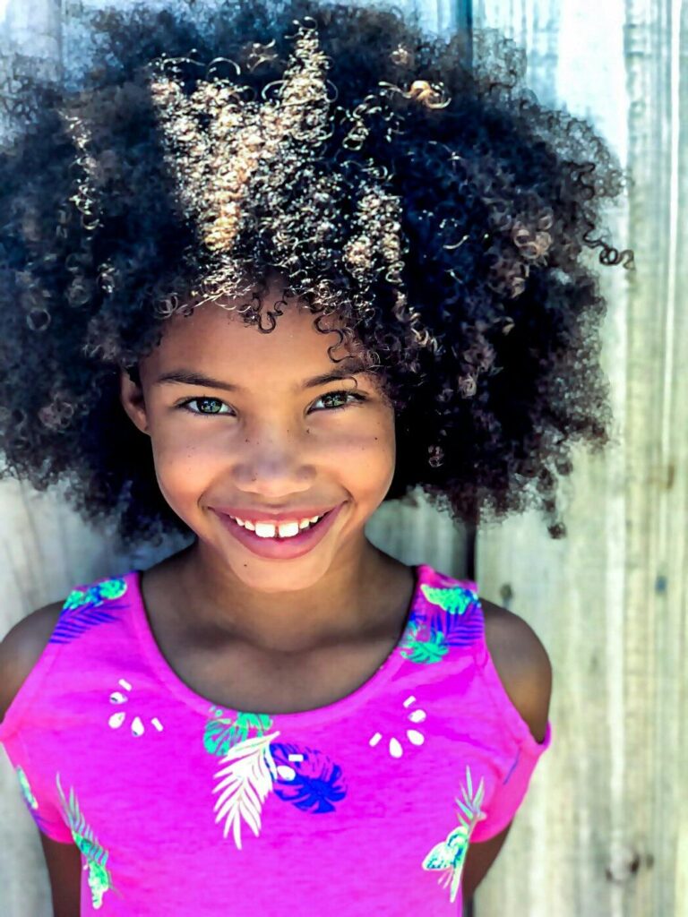 A joyful young girl showcases her beautiful curly Afro hair and bright smile in an outdoor portrait against a wooden backdrop.